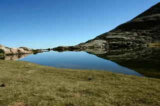 Calm high-altitude lake surrounded by rocky terrain and alpine grass, reflecting the landscape under a clear sky.