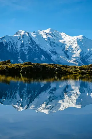 Snow-covered Mont Blanc reflected in a calm alpine lake under a clear blue sky.