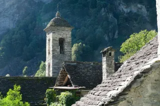 Stone rooftops and a bell tower of a traditional alpine village nestled in a green mountain valley.